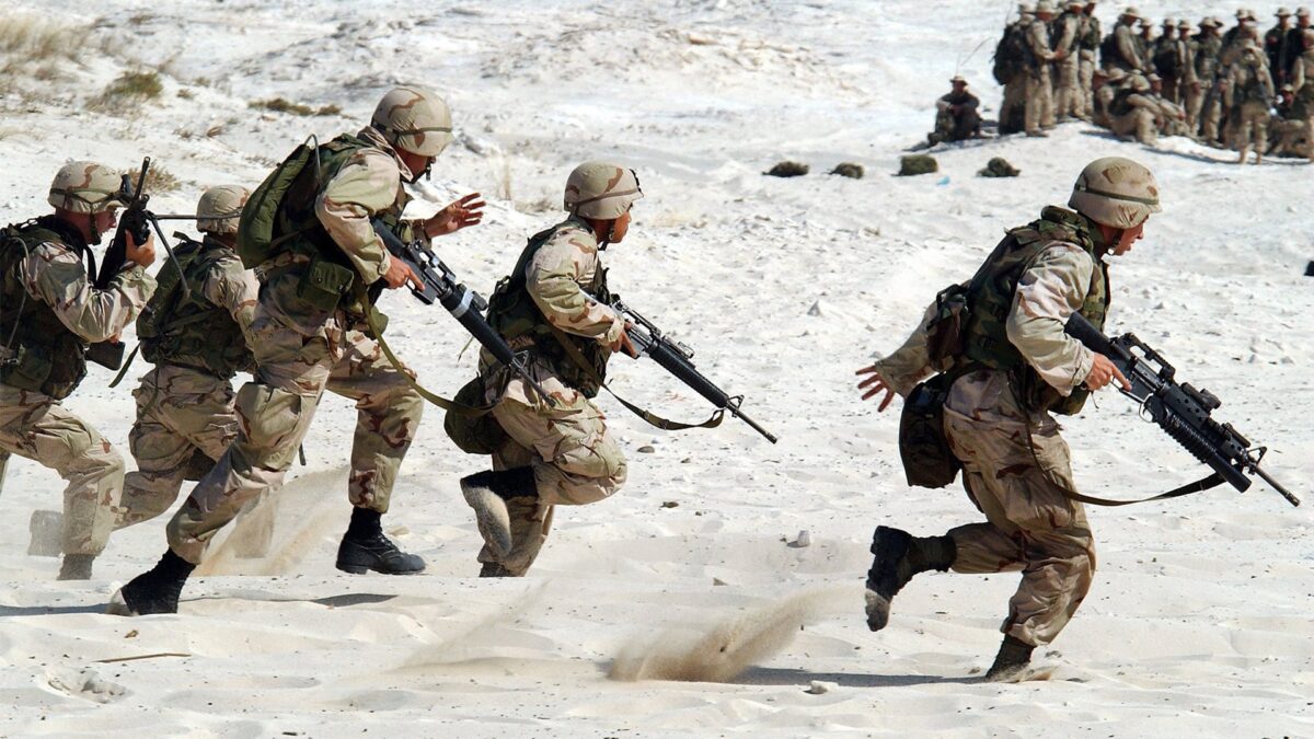 Armed soldiers run across a sandy landscape in an exercise.
