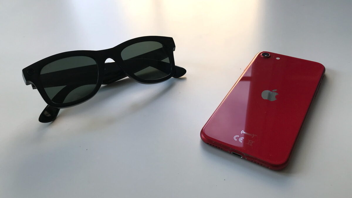 A Ray-Ban Stories lies next to a red iPhone on a table.