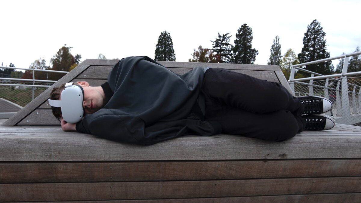 A young man lies covered on an outdoor bench with Meta Quest 2 on his face.