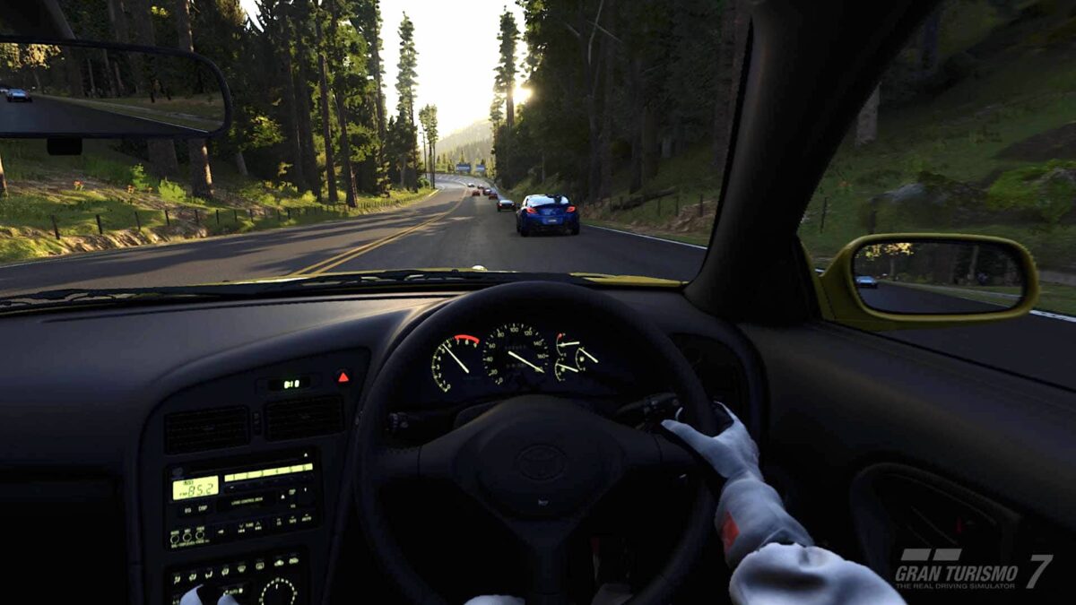 View from the cockpit of a GT7 race car onto a road bordered by forest on both sides.