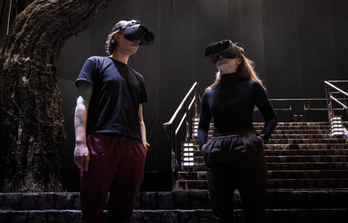A man and a woman wearing Varjo VR headsets stand on the stage of the Finnish National Opera, where an old tree is placed on a staircase.