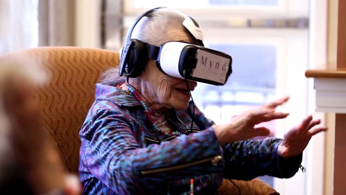 An elder woman sits on a chair wearing a VR headset.