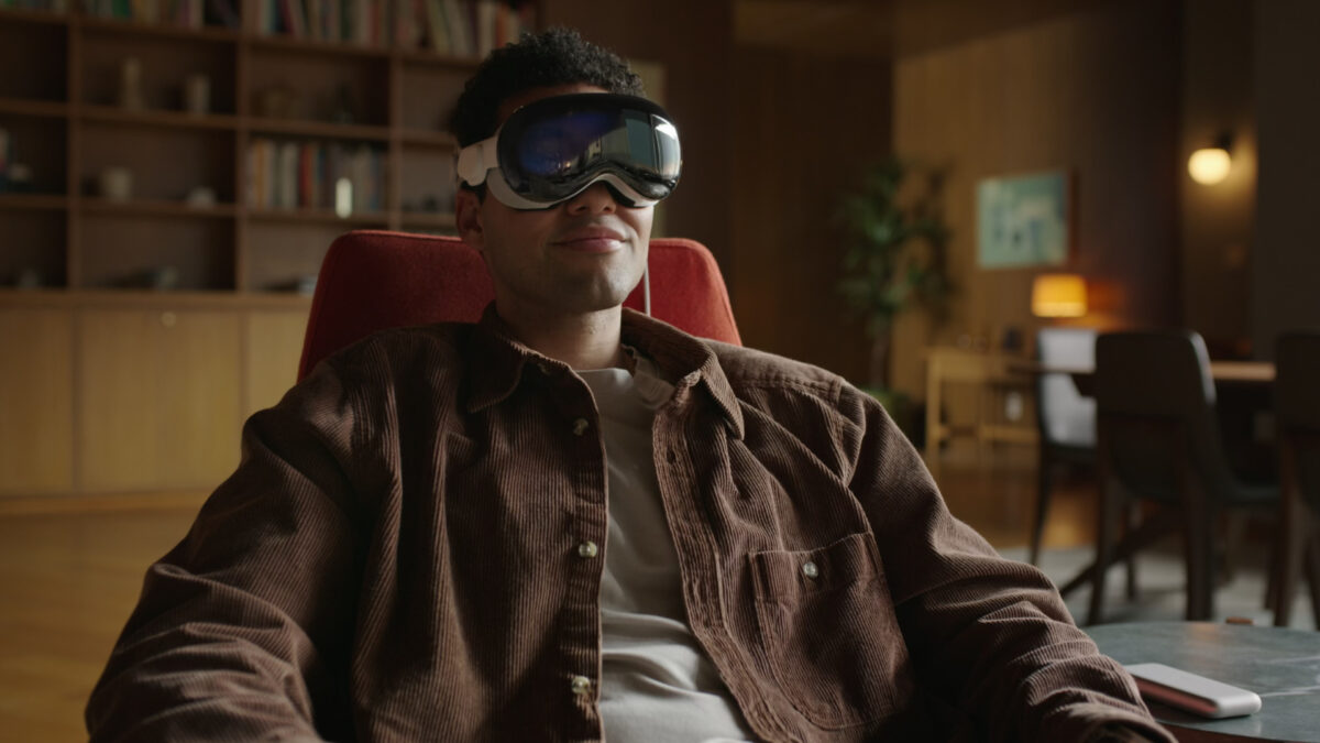 A man sits on an armchair with a grin on his face and the Apple Vision Pro on his head.