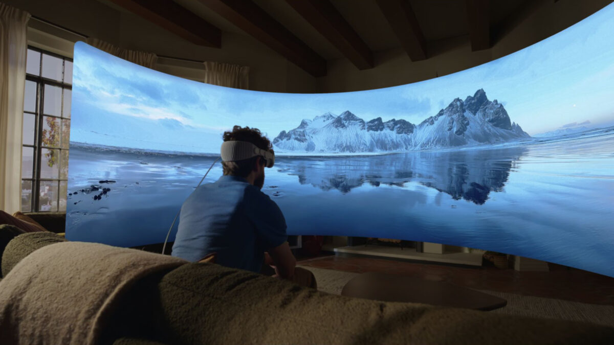 A man sits in his living room with an Apple Vision Pro on his head, looking at a virtual screen showing a landscape image.