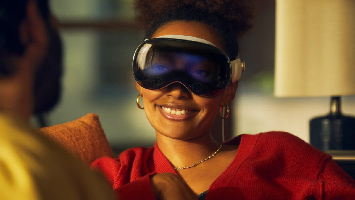 Woman wears Apple Vision Pro on a couch and is facing a person smiling. Her eyes shine through the surface of the headset.
