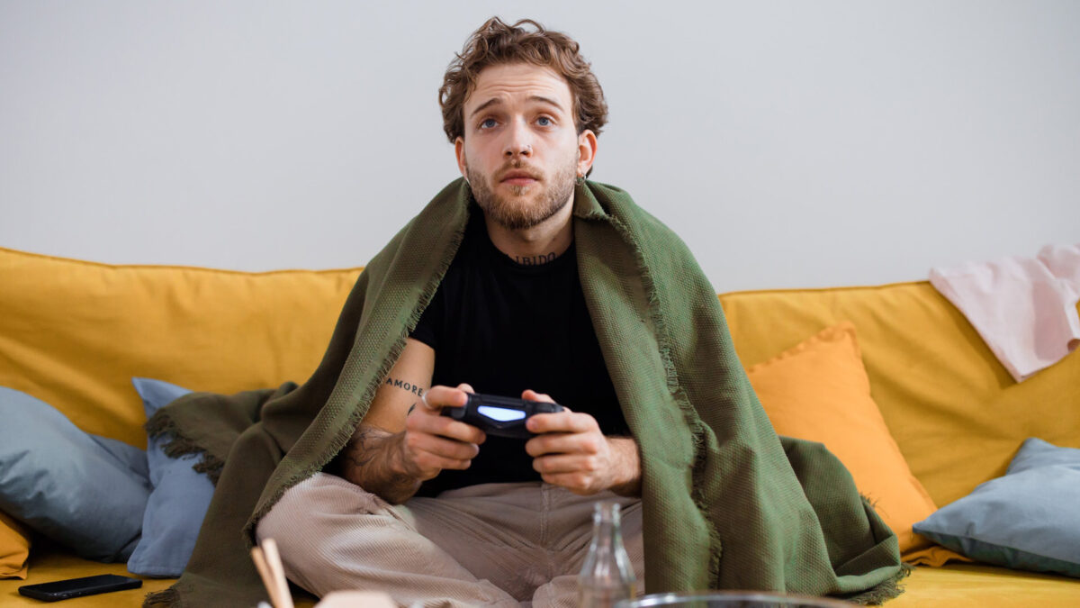 A young man sits on a couch wrapped in a blanket and holds a gamepad in his hand.