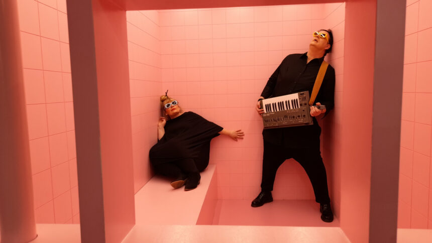 A man and a woman in black robes huddle in the corners of a pink room.