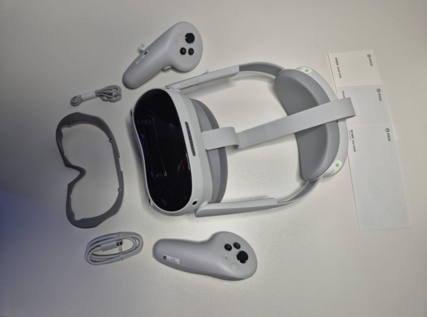 The Pico 4 Ultra VR headset lies on a white table with two VR controllers, a headset spacer, a USB-C cable and three small booklets.