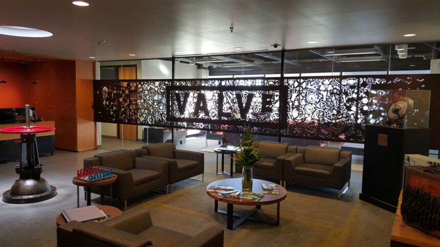 Modern office lounge with brown leather seating, backlit Valve logo art panel, chess set, and industrial-style decor against orange and white walls.